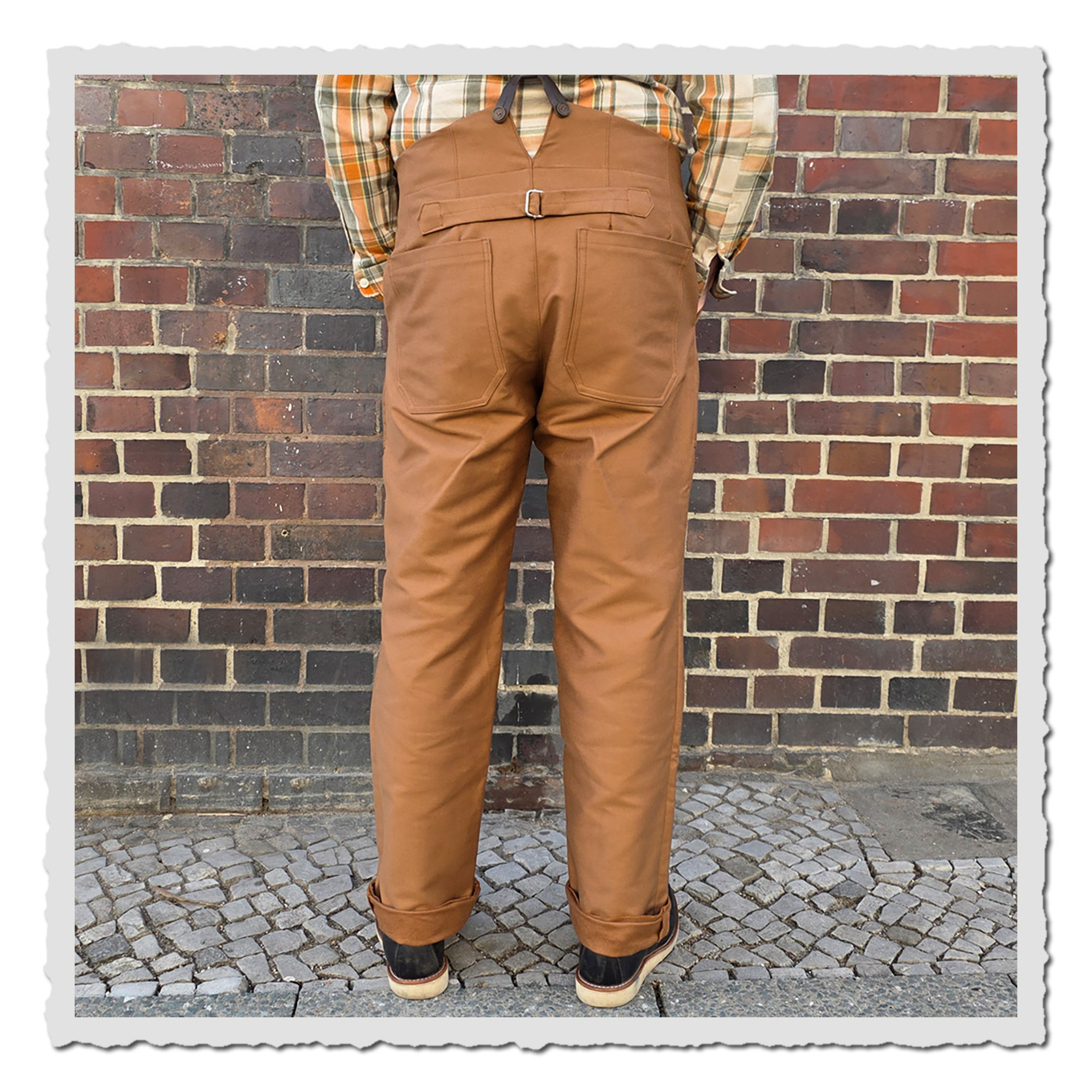 Rückansicht: braune Arbeiterhose am Körper getragen, vor Ziegelmauer, Beine und Schuhe sichtbar.